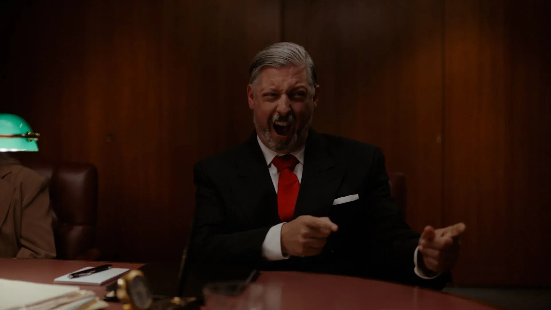 A Big Paper executive in a dark suit and red tie laughing in a wood-paneled boardroom